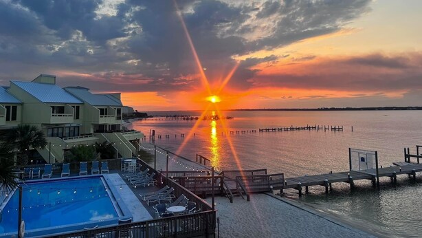 A sunset over a calm body of water with a dock extending into the water. There is a pool area with lounge chairs and a building with a sloped roof in the foreground.