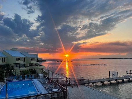 A sunset over a calm body of water with a dock extending into the water. There is a pool area with lounge chairs and a building with a sloped roof in the foreground.