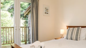 Une chambre avec un lit en bois, du linge de lit blanc, une table de chevet avec une lampe, un tableau encadré au mur et un balcon donnant sur un paysage verdoyant.
