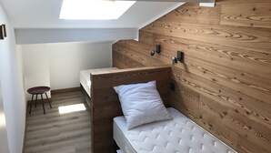 Una habitación compacta con techo inclinado, paneles de madera, una litera, un taburete pequeño y un tragaluz.
