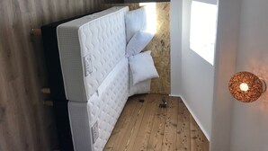 Una habitación compacta con piso de madera, un colchón blanco con almohadas, una mesita y una lámpara fijada a la pared.