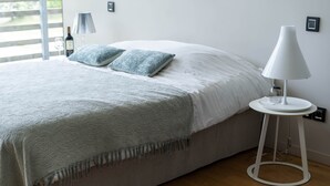 Ein ordentlich bezogenes Bett mit grau-weißer Decke, zwei Kissen und einer weißen Nachttischlampe auf einem kleinen Tisch daneben.