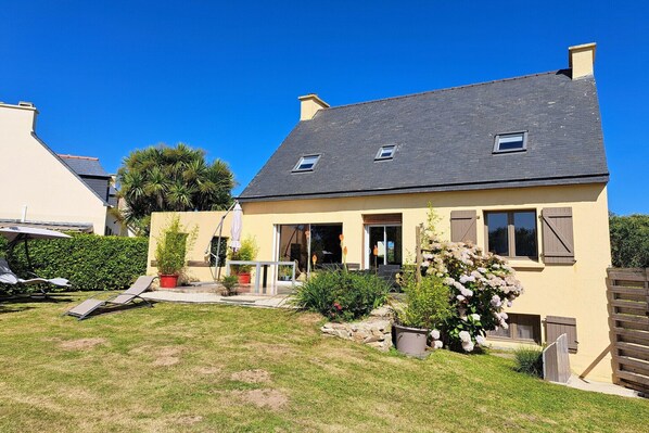 Une maison à deux étages, dotée d’un toit en pente, d’un petit porche à l’avant et d’un jardin bien entretenu avec des plantes en pot et une pelouse.