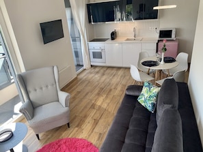 Ein kompakter Wohnbereich mit Sofa, Zähneform-Ohrensessel und Esstisch mit Stühlen. Ein Küchenbereich ist erkennbar, mit Mikrowelle, Toaster und Spüle.