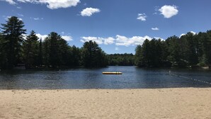 En strand med en sandstrand, en lugn sjö och en rad träd i bakgrunden.