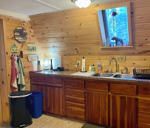 Interior de una cabaña de madera con cocina, que incluye un lavamanos, armarios y una ventana con un adorno.