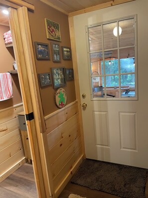 Een houten interieur met een deur, foto's aan de muur, een klein vloerkleed en een raam met uitzicht op bomen.