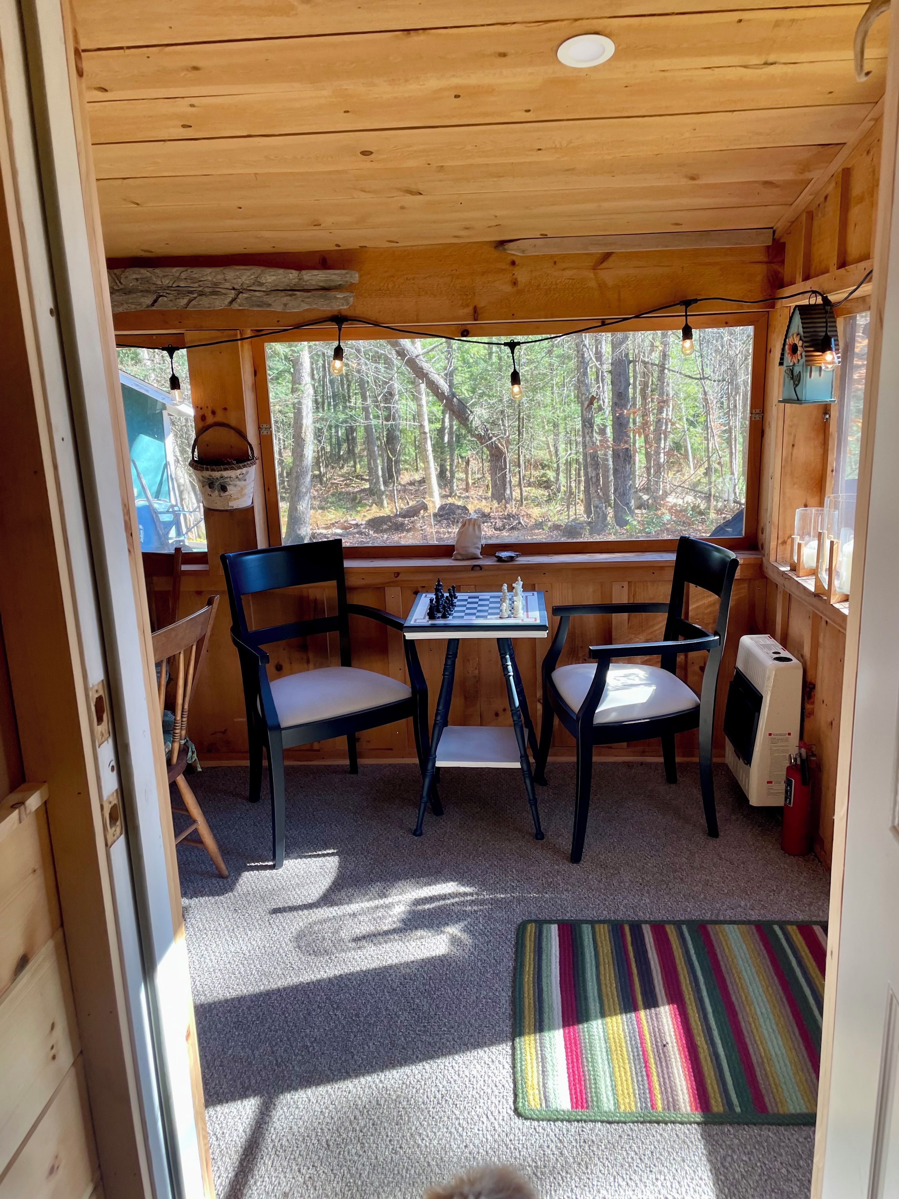 L'interno accogliente di una baita, con un soffitto in legno, una scacchiera su un tavolino, due poltrone, un tappeto colorato e una finestra con vista su una foresta.