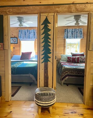 Een blokhutinterieur met twee slaapkamers, elk met een bed, een raam met gordijnen en een plafondventilator.