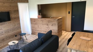 A modern living space with a wooden wall, a dark sofa, a small round table, and a wooden dining table. - 4-6 Pers apartment in the heart of the Chartreuse at the foot of the ski slopes (Sarcenas)