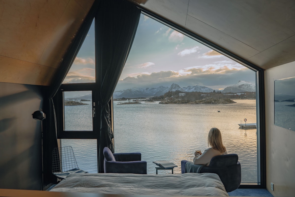 A room with a large window overlooking a lake and mountains, a bed, a chair, and a table.