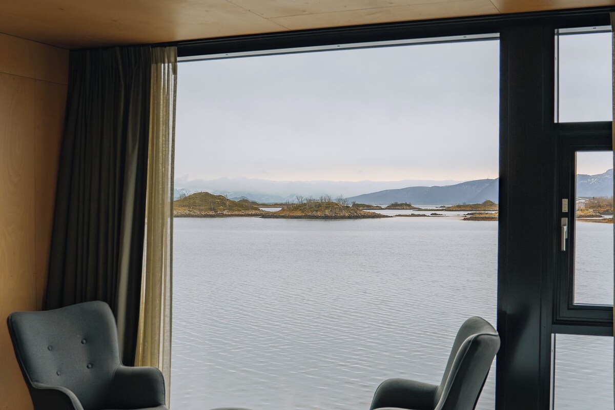Una habitación con una cama, dos sillones y un ventanal con vistas a un lago y montañas.