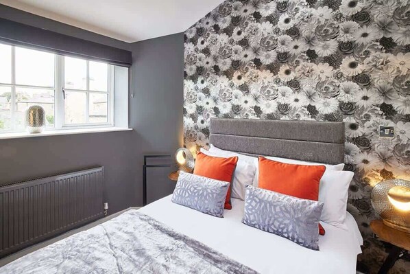 Une chambre à coucher avec une tête de lit grise, des coussins blancs et orange, une couvre-lit à motifs et un papier peint à fleurs.