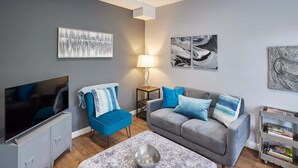 Uma sala de estar moderna com um sofá cinzento, uma poltrona azul, uma TV de tela plana e obras de arte nas paredes.