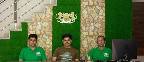 Three individuals seated at a reception desk with a stone wall and green carpeted steps in the background.