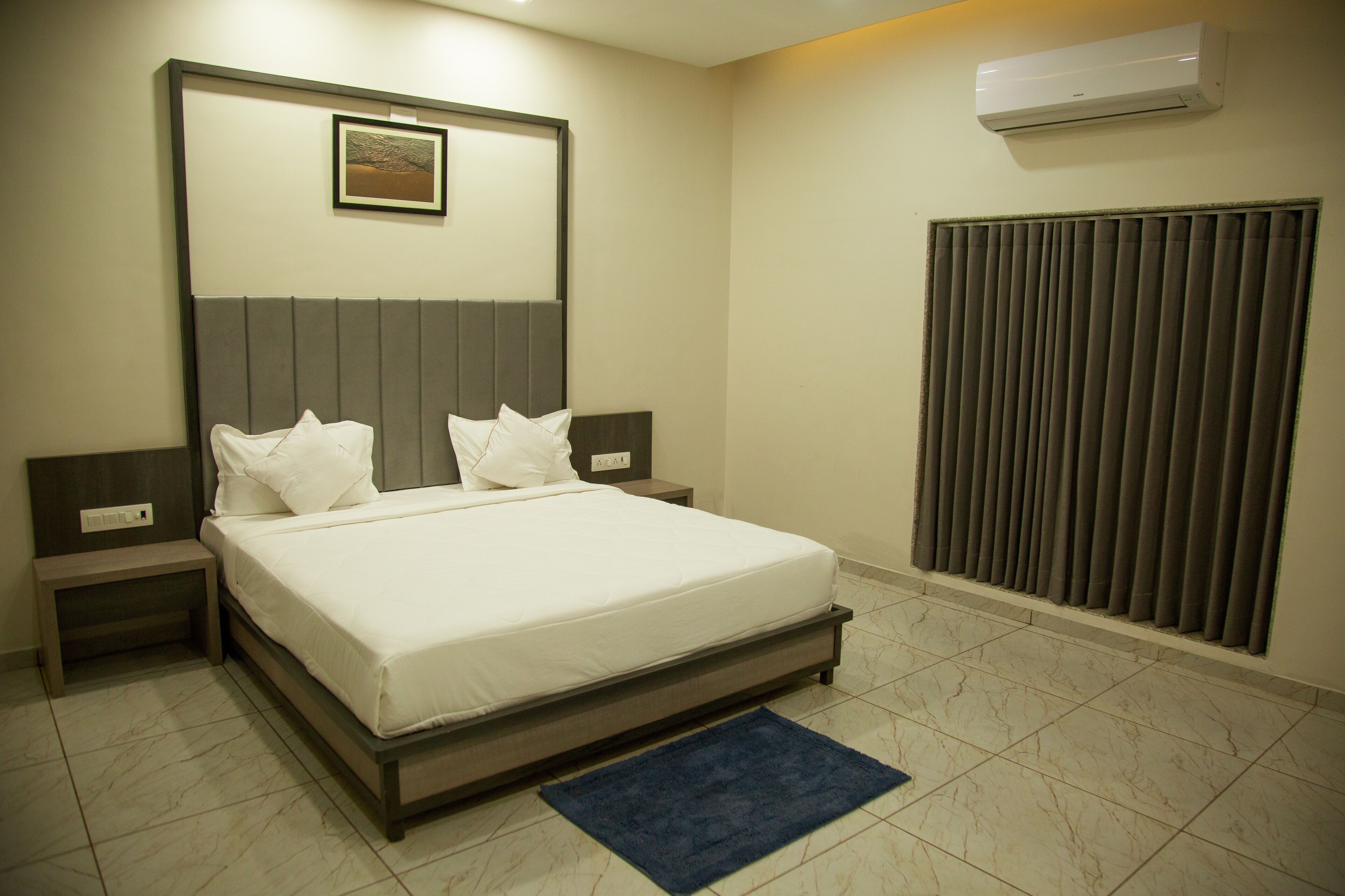 Une chambre à coucher bien rangée, avec un lit, des tables de chevet, une petite table, un tapis bleu, un climatiseur fixé au mur et une fenêtre avec des stores.