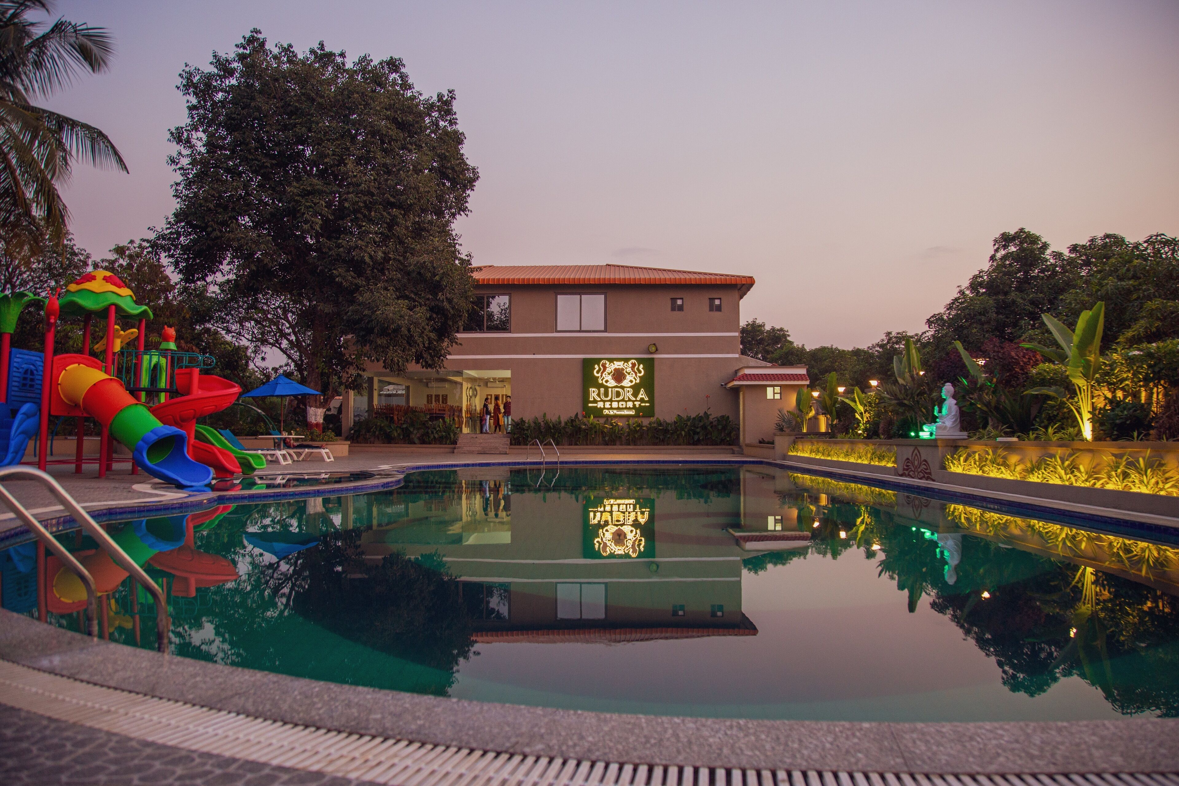 A swimming pool with a slide, surrounded by trees and a building with a sign.