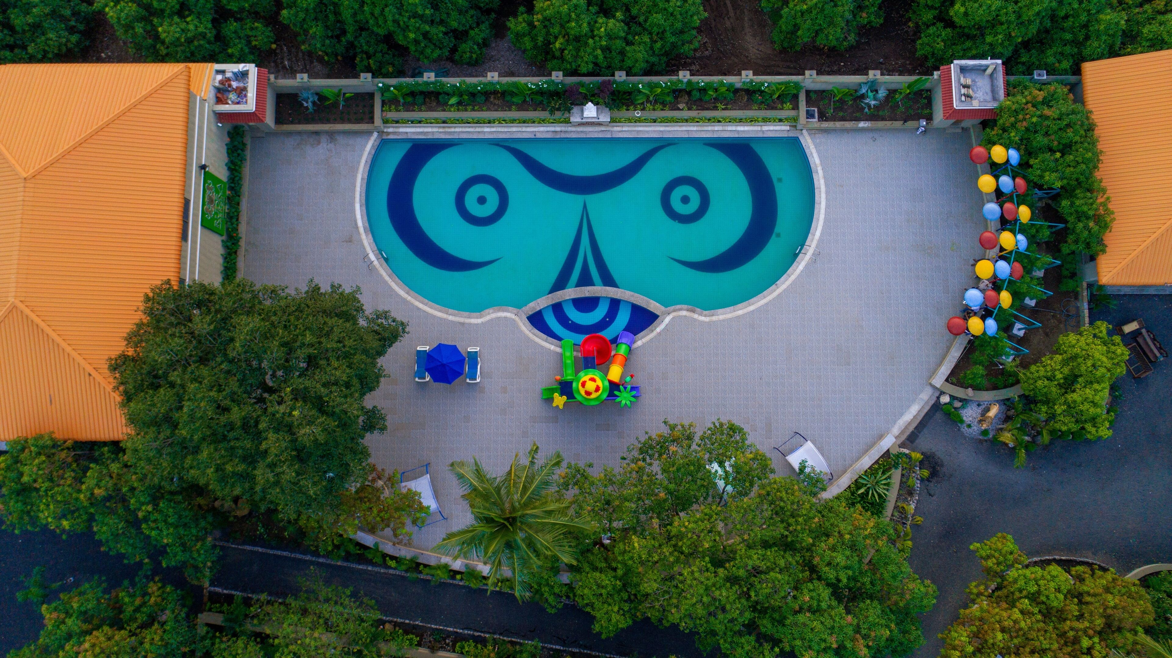 An aerial view of a swimming pool with a unique design, surrounded by greenery and a small seating area.