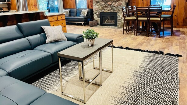 A cozy living room with a stone fireplace, wooden walls, a sofa, a dining table with chairs, and a kitchen area with a microwave and refrigerator. - Lazy Bear Bungalow Pet Friendly Lake Escape  (Truckee)