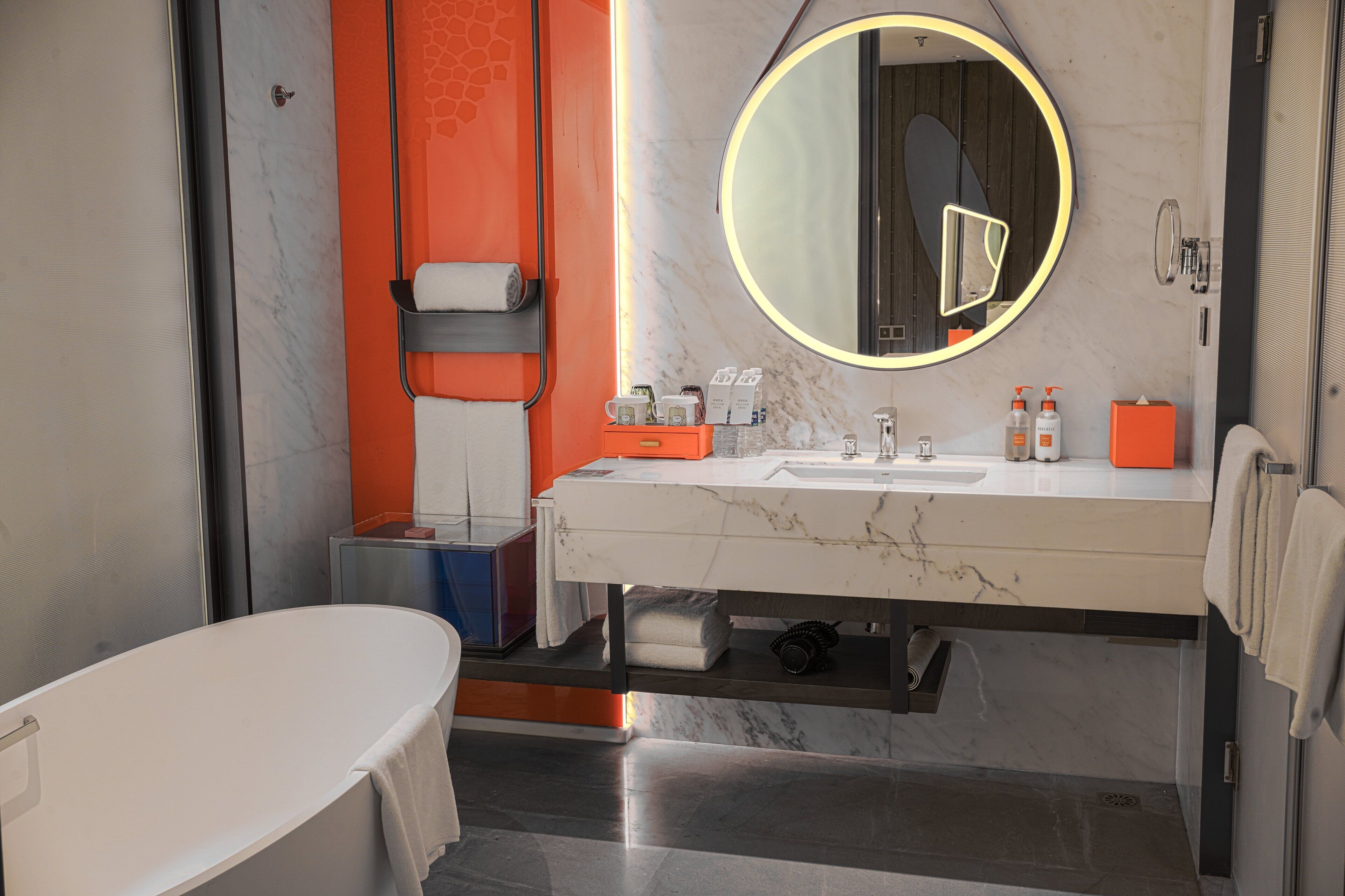 Un baño moderno con lavamanos de mármol, un espejo grande con luz circular y una bañera independiente.