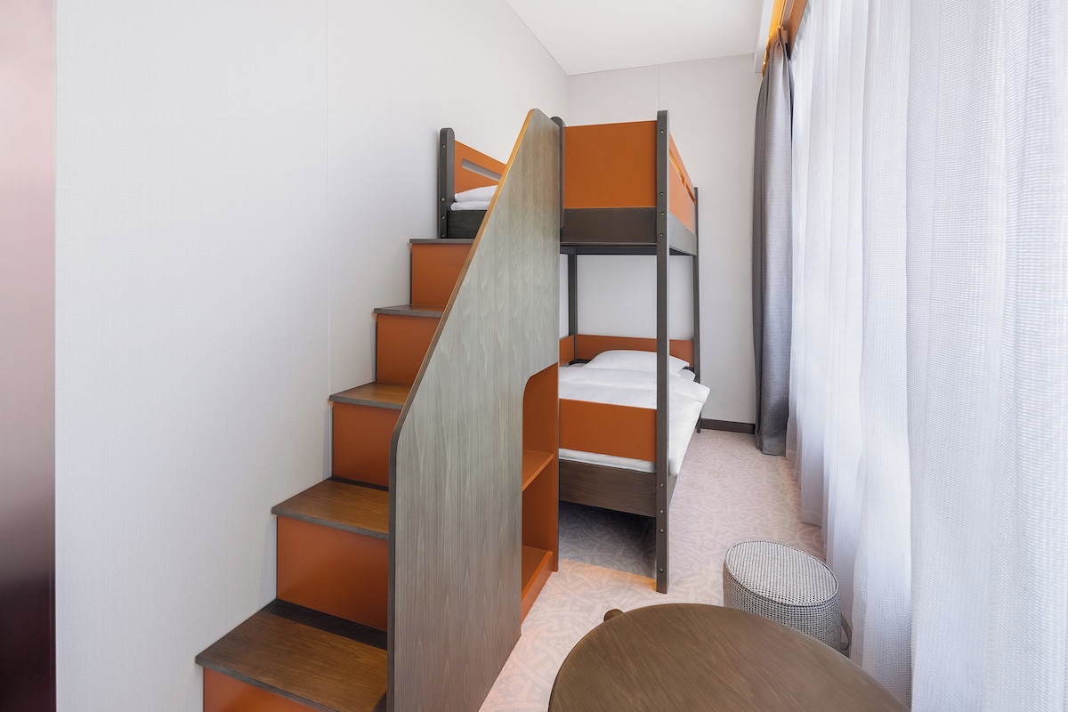 Ein kleines Zimmer mit einem Stockbett, einem kleinen runden Tisch und einem Korbstuhl.