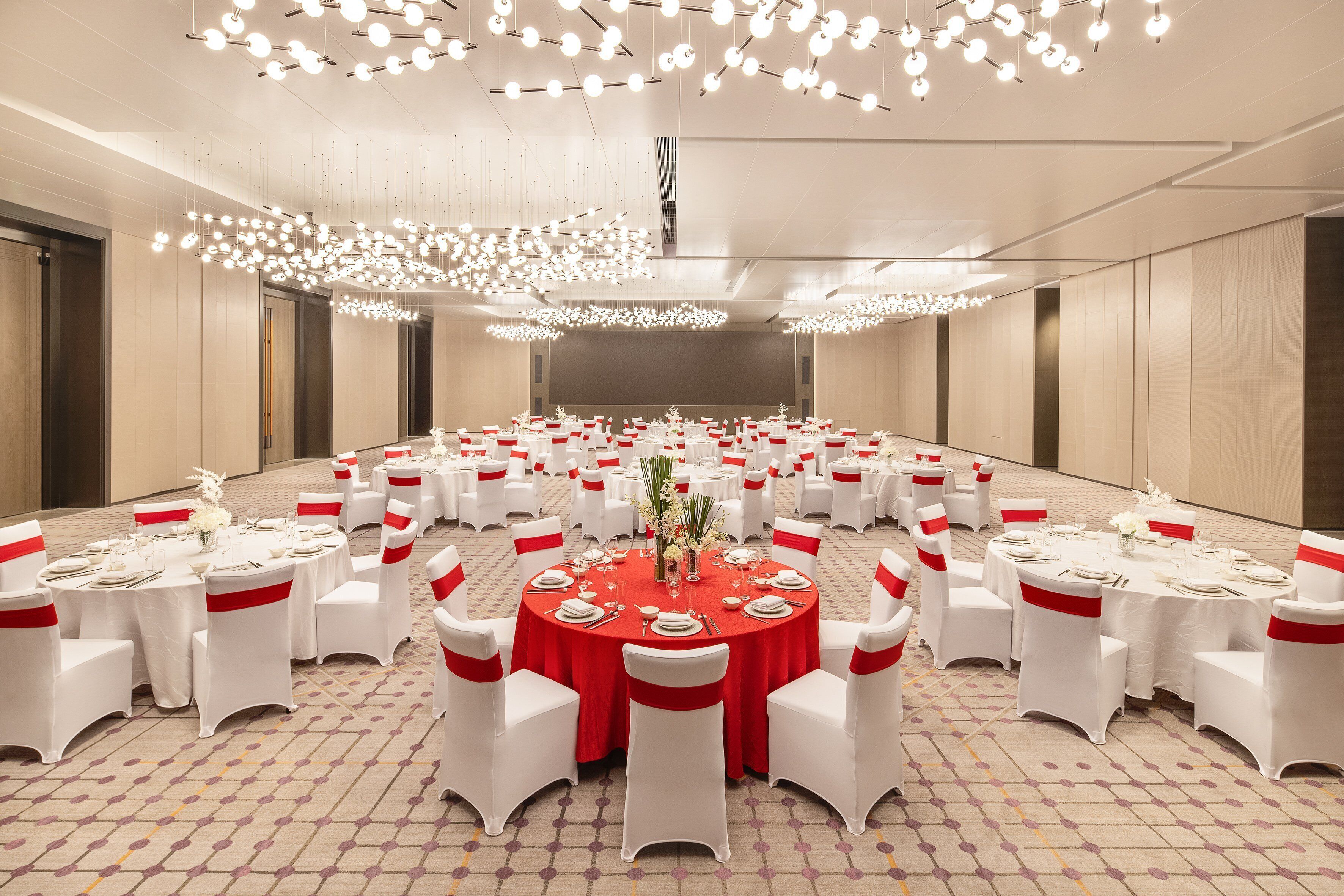 Une grande salle de conférence avec des tables rondes, des chaises blanches et des rubans rouges. Les tables sont dressées avec des nappes blanches, des assiettes et de la verrerie. La salle a un plafond haut et est éclairée par des luminaires suspendus.