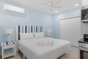 A hotel room with a bed, bedside tables with lamps, a ceiling fan, an air conditioning unit, a wardrobe, and a kitchenette.