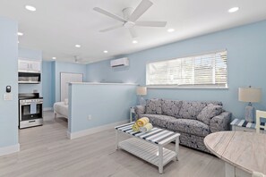 Ein modernes Wohnzimmer mit einer Couch, einem Couchtisch, einem Deckenventilator und einer Küchenzeile mit Backofen und Schränken.