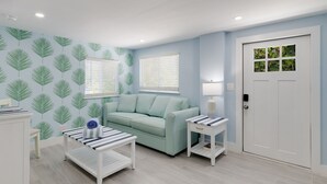 Una habitación con pared azul claro, papel tapiz estampado con hojas verdes, sofá verde claro, puerta blanca con ventanal y mesa con cojines a rayas.