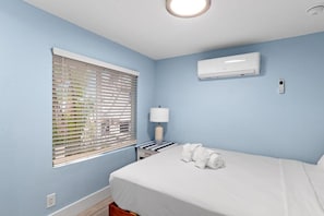 Ein Hotelzimmer mit Bett, Fenster mit Jalousien, Klimaanlage, Nachttischlampe und Steckdose.