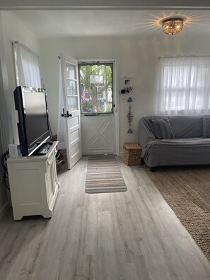Une pièce avec un téléviseur posé sur un meuble, un canapé gris, une porte donnant sur l’extérieur et un petit tapis sur le sol.