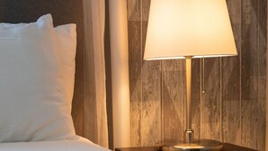 Ein ordentlich bezogenes Bett mit weißen Leinen und einem einzelnen weißen Kissen, ein Nachttisch mit einer Lampe und eine holzstrukturierte Wand im Hintergrund.