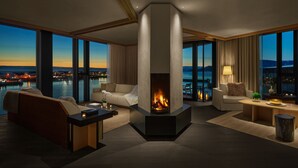 Una sala de estar moderna con chimenea, ventanas amplias y vista a un paisaje urbano al anochecer.