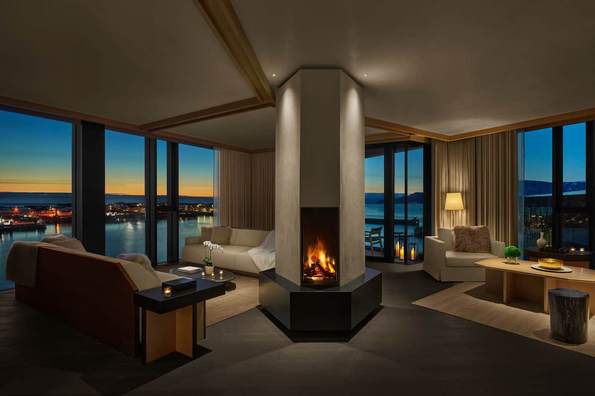 A modern living room with a fireplace, large windows, and a view of a cityscape at dusk.