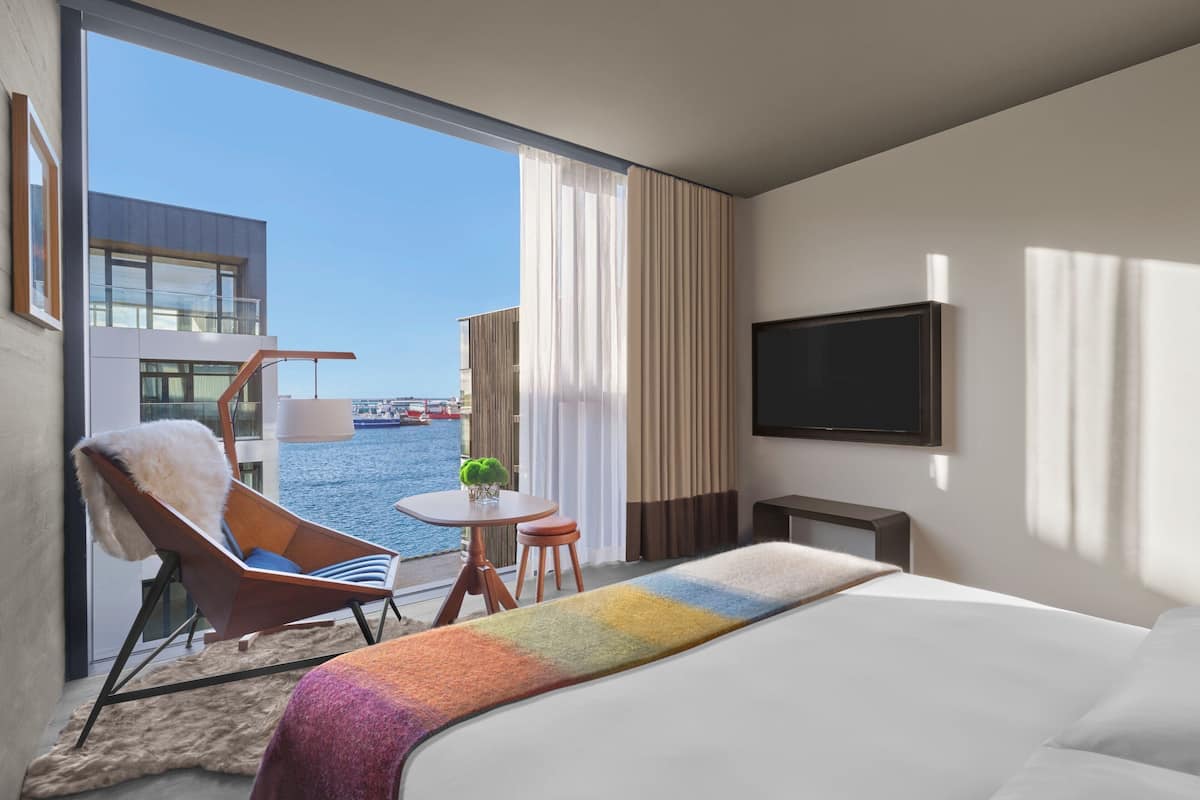 A hotel room with a large bed, a TV, a chair, a small table, and a view of the water and buildings.