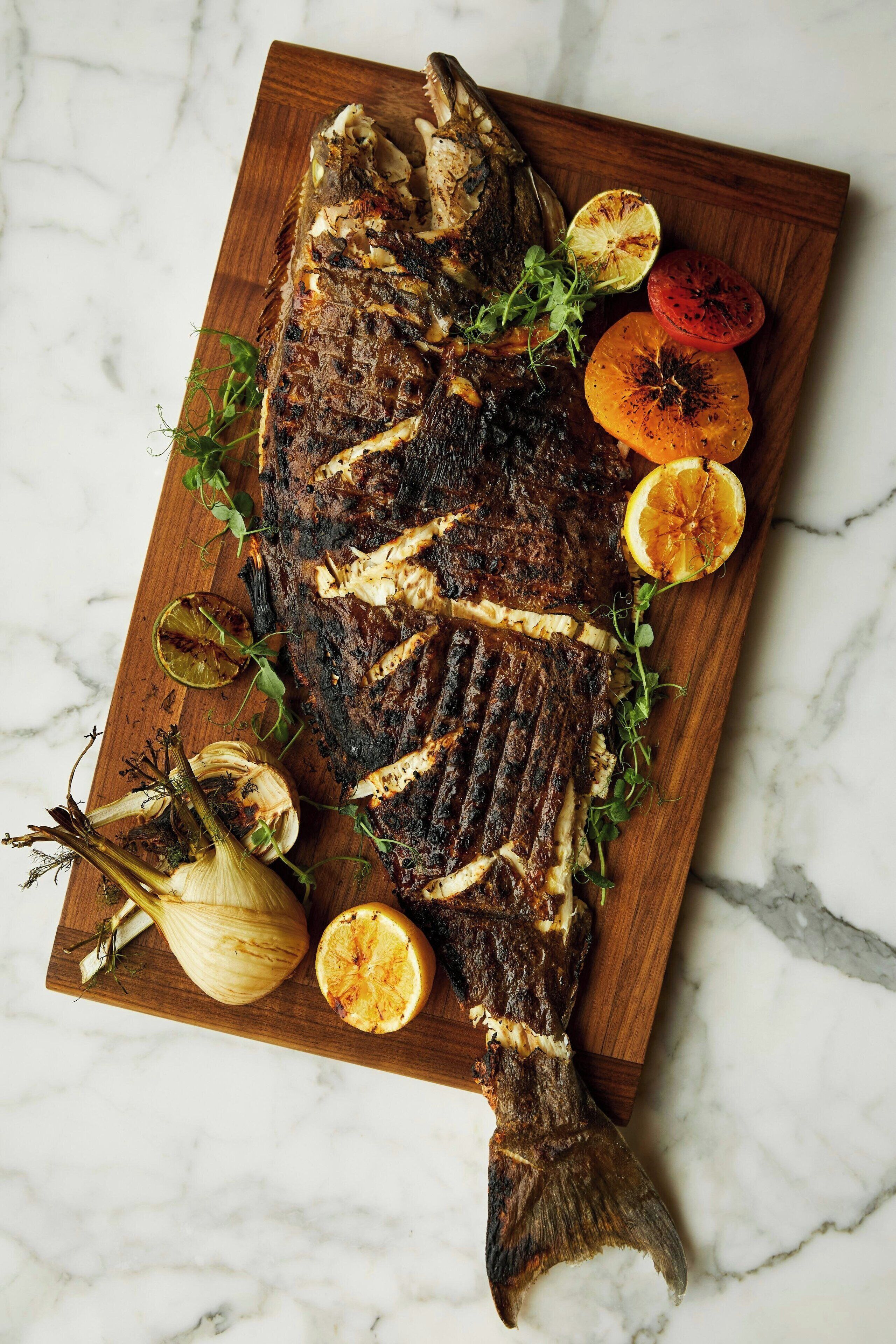 A grilled fish on a wooden board with lemon slices and herbs.