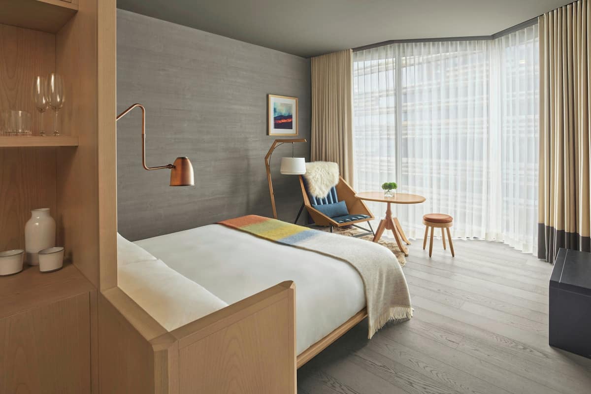 A hotel room with a wooden bed, a desk with a chair, a lamp, a painting on the wall, and a large window with curtains.