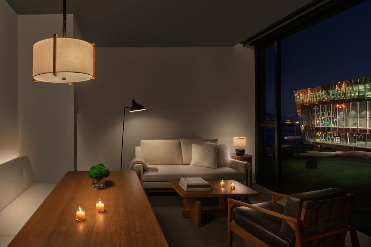 A modern living room with a sofa, coffee table, and a dining table with candles. A lamp and a view of a building are also visible.