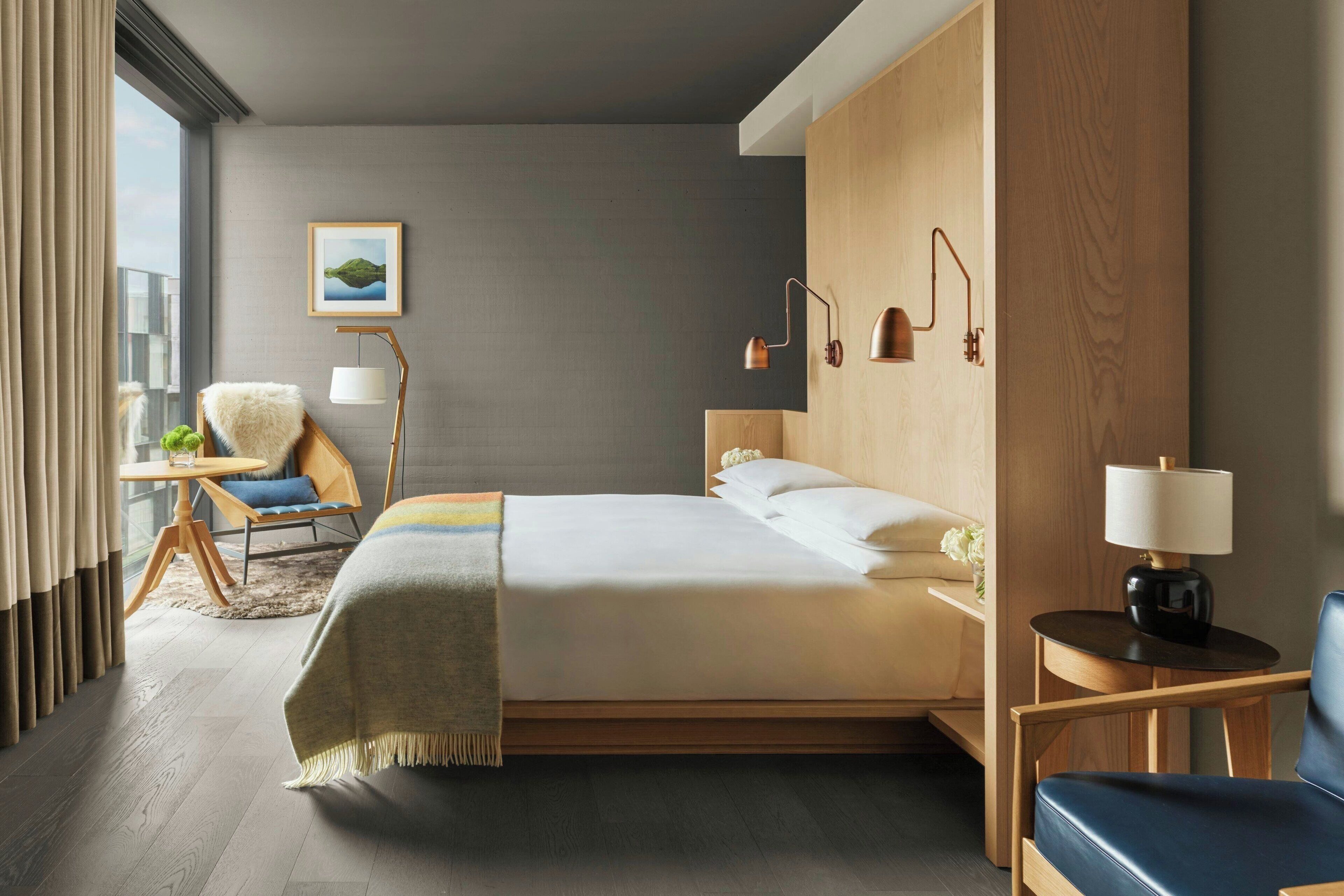 Una camera d'albergo moderna con un letto grande, una testiera in legno, un comodino con una lampada, una sedia e vista sulla città attraverso ampie finestre.
