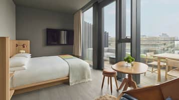 A modern hotel room with a large bed, a small round table, and a view of the city through large windows.