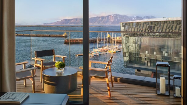 Un balcón con muebles de madera, una mesa redonda y vistas a un puerto con barcos y montañas al fondo.