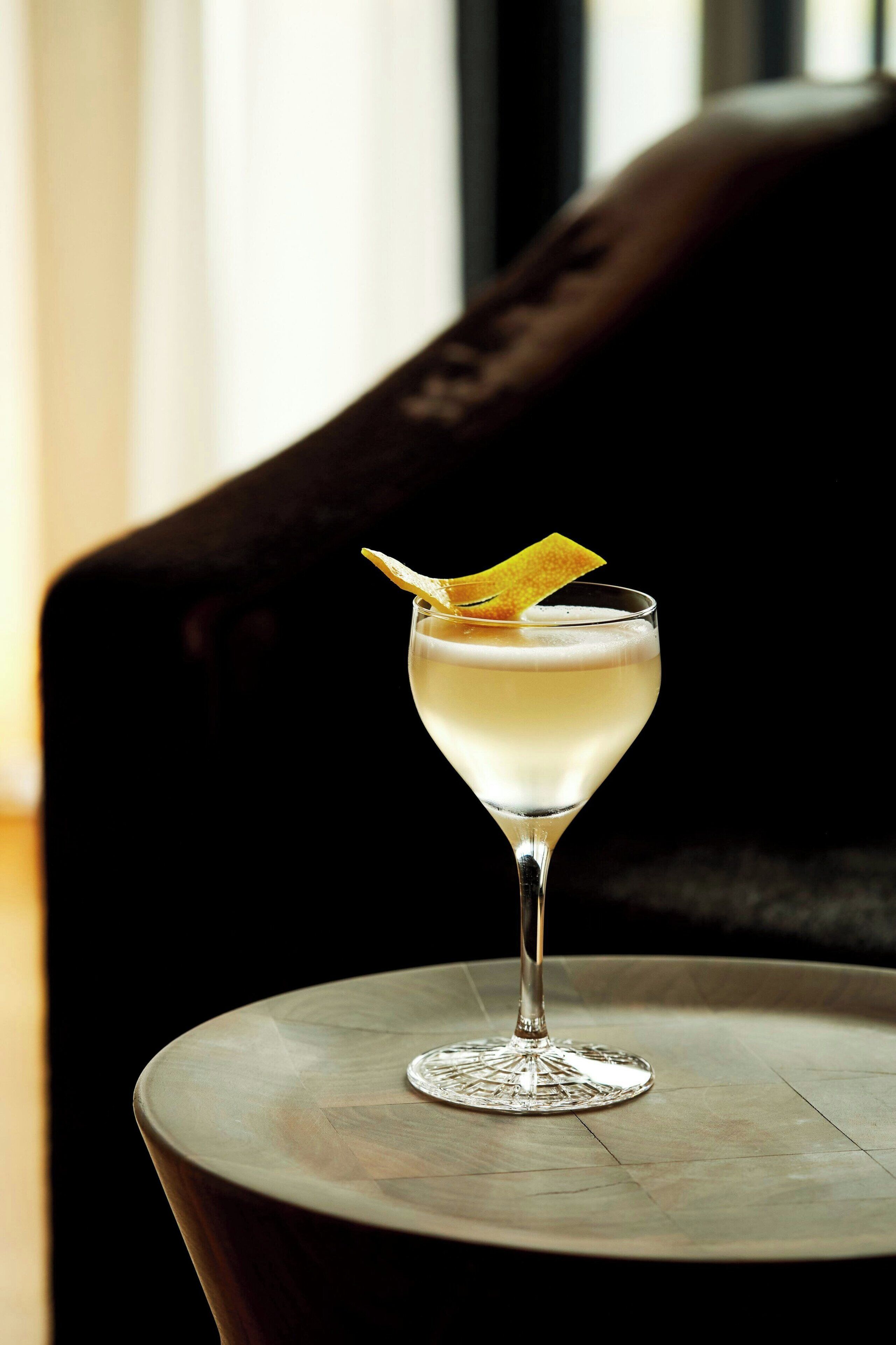 Un verre à cocktail sur une table ronde, avec une chaise à l’assise rembourrée de couleur sombre en arrière-plan.