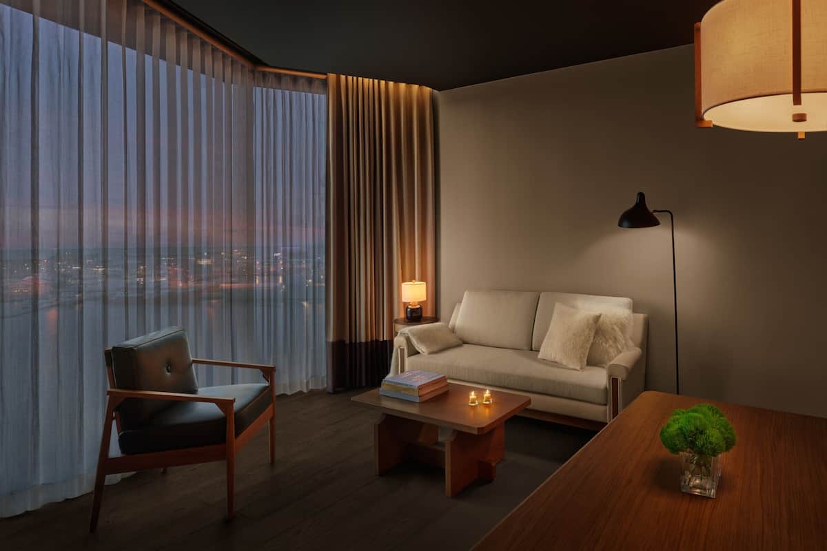 A modern hotel room with a large window, a sofa, a chair, a coffee table, and a lamp.