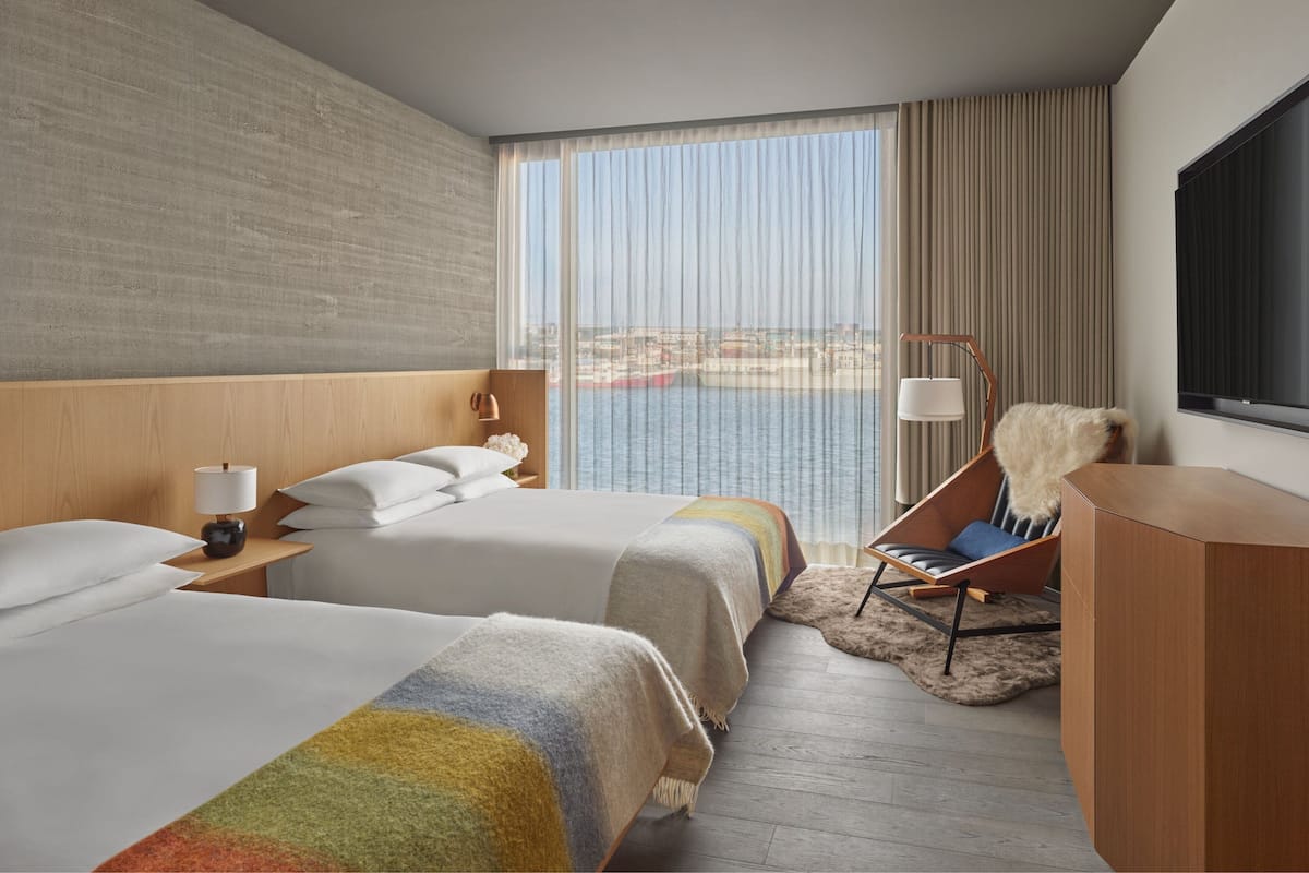 A hotel room with two beds, a large window with a view of the sea, a chair, and a desk.