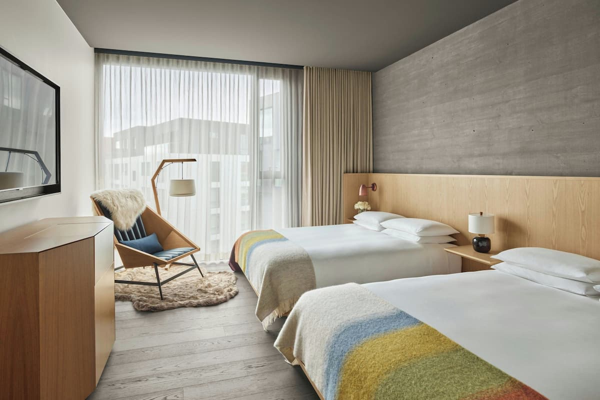 A hotel room with two beds, a wooden headboard, a large window with sheer curtains, a chair with a fur throw, and a desk with a lamp.