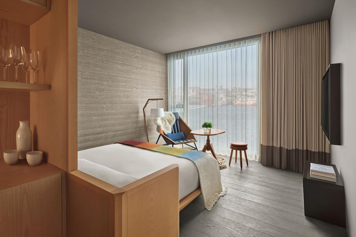 A hotel room with a bed, a chair, a small table, a TV, and a view of the city through large windows.