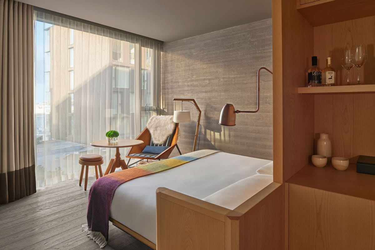 A modern hotel room with a large bed, a chair, a small table with a lamp, and a view of the city through large windows.