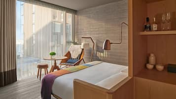 A modern hotel room with a large bed, a chair, a small table with a lamp, and a view of the city through large windows.