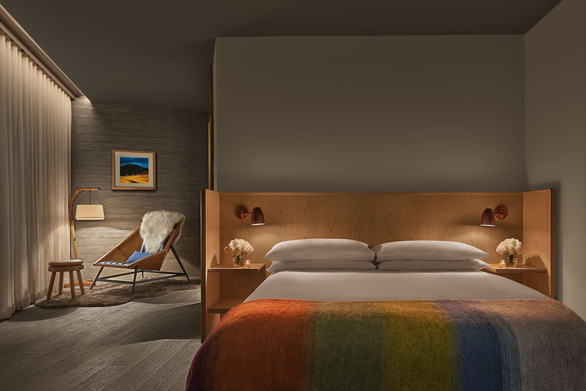 A modern hotel room with a large bed, a wooden headboard, two bedside lamps, a chair with a fur throw, and a framed picture on the wall.