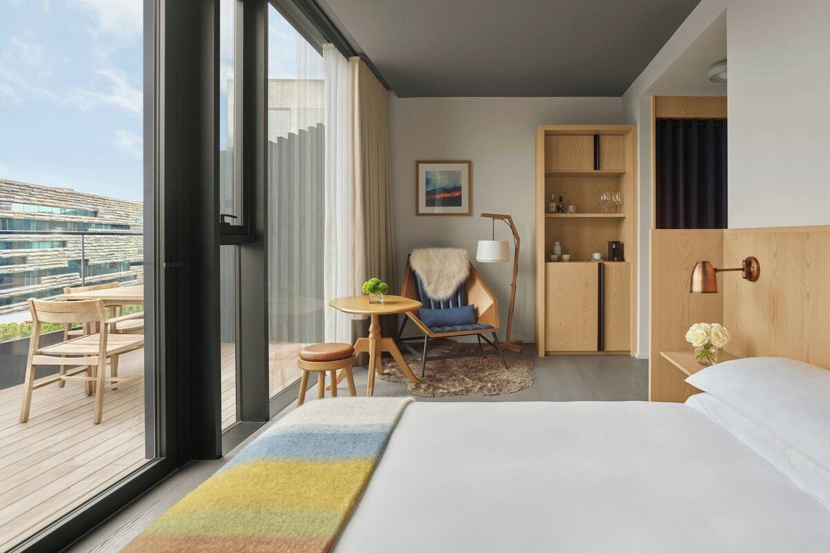 A hotel room with a large bed, a wooden desk, a chair, a small table with a lamp, and a view of the city through large windows.